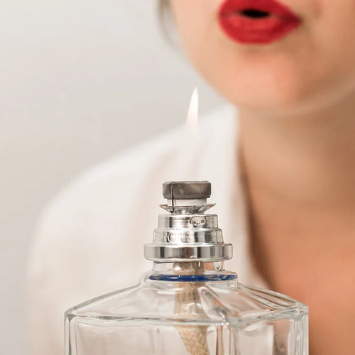 A close-up of a clear glass perfume bottle with a lit silver and black wick igniter, set against a soft-focus background of a woman in a white shirt and red lipstick.
