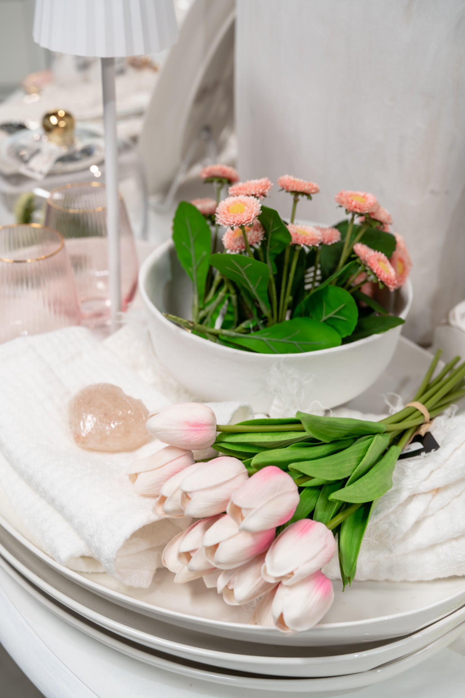 A styled tabletop arrangement featuring a white ceramic bowl filled with pink daisies and green stems, alongside a bouquet of pale pink tulips on white folded towels, with a softly blurred home decor background.