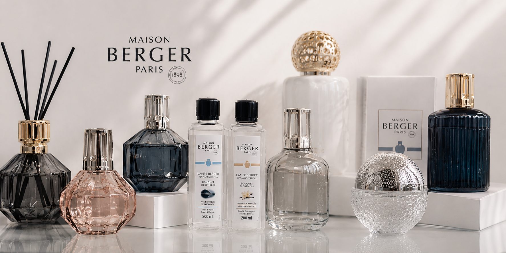A collection of Maison Berger Paris home fragrance products, including a reed diffuser, a pink faceted bottle, a dark blue glass bottle, two white spray bottles, a clear glass bottle, and a textured silver orb, arranged on white pedestals against a light background with soft shadows.